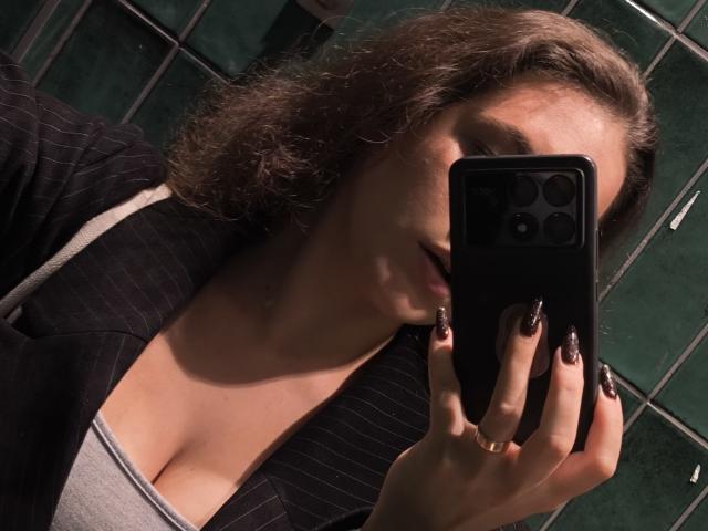 Dreia, met krullend bruin haar, een gestreept jasje en een grijze top, houdt een zwarte telefoon vast, en laat haar donker geverfde nagels en decolleté zien, in een slecht verlichte badkamer..