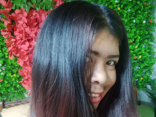 MariahSquirting69 has long, shiny black hair covering one eye, looking at the camera with a slight smile, showing red lipstick and a small beauty mark on her nose.