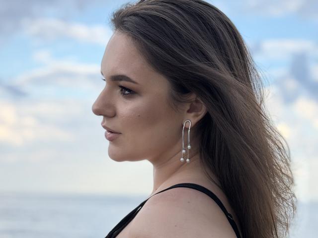 Dreia's side profile, showing her brown eyes, subtle makeup, and long brown hair. She's wearing a black top and silver, curved, dangling earrings with small pearls.