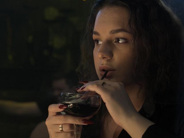 Dreia, avec ses cheveux noirs et bouclés, sirote du vin rouge dans un verre muni d'une paille noire, et regarde sur le côté. Elle a les ongles rouges et porte une bague en or à la main gauche..