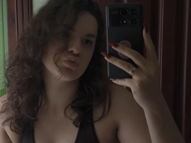 Dreia, met krullend bruin haar, een donkere tanktop en rode nagels, houdt een zwarte telefoon vast en maakt een spiegelzelfportret, waarbij haar gezicht en bovenlichaam te zien zijn. Haar borsten zijn lichtelijk zichtbaar..