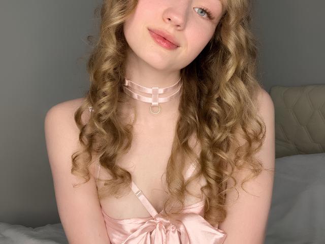 BulahLiebman, con i capelli biondi ricci, una camicetta di seta rosa e una collana choker rosa, mostra piccoli seni, pelle chiara e labbra rosa, sorridendo leggermente. Si trova in una camera da letto illuminata da una luce soffusa..