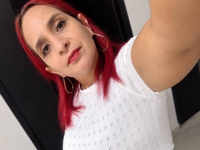 KateMonroe with red hair, white eyelet top, small hoop earrings, and pink lipstick, taking a selfie, arm raised, smiling slightly, fair skin, brown eyes, cute face, medium breasts.