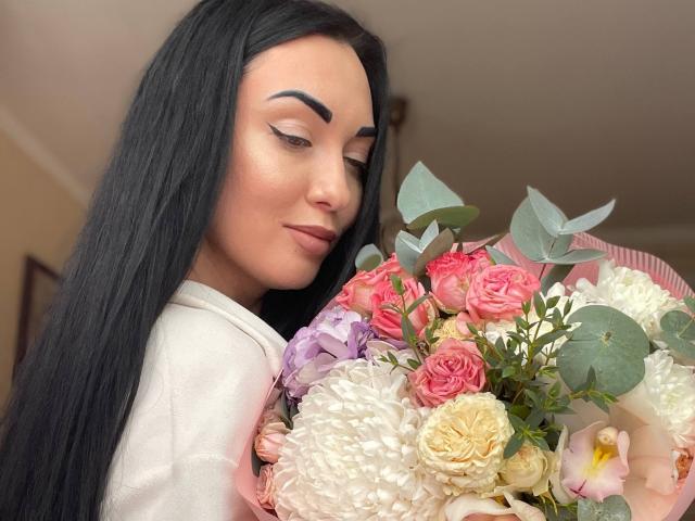 Xioni with long black hair, holding a colorful bouquet of roses, carnations, and eucalyptus. She's wearing a white top, looking down with a subtle smile.