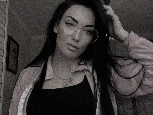 Xioni with long black hair, glasses, and pink jacket over black top, hand in hair, slight smirk, gray-scale photo, small necklace, subtle makeup, relaxed, confident.