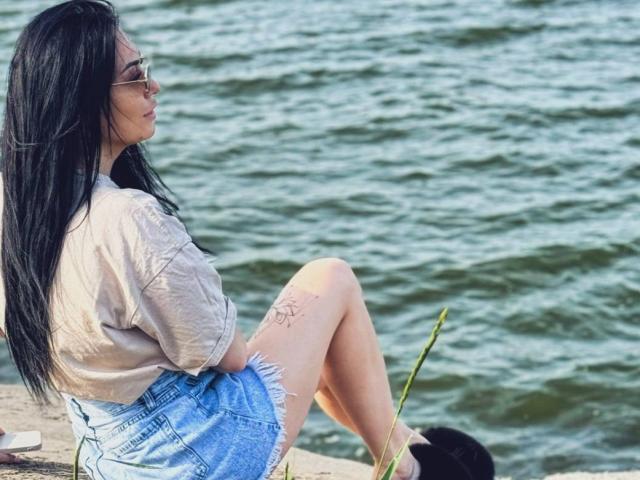 Xioni with long black hair, glasses, beige shirt, and blue denim shorts, sitting by the water, legs crossed, showing a tattoo on her thigh, holding a green stem.