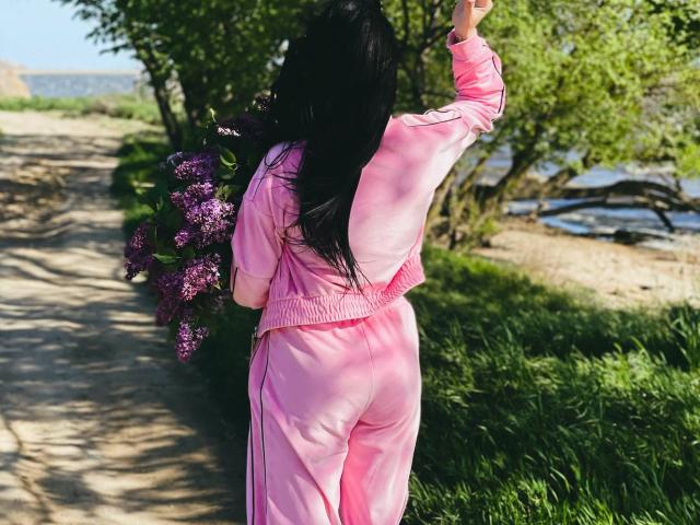 Xioni in pink track suit, facing away, with black hair, raising right arm, standing on path with purple lilacs, showing her ass, near grass and trees.