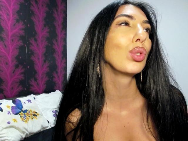 BrunetteBabe69 with long black hair, pouty pink lips, and shiny skin, wearing hoop earrings, looking up with a slightly open mouth, showing a bit of cleavage.
