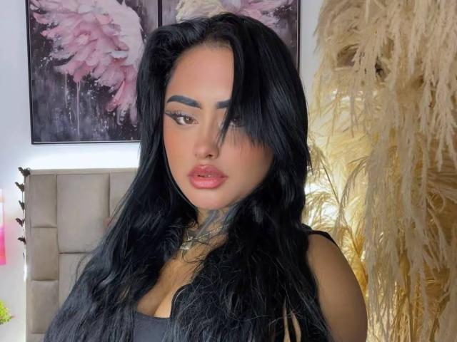 LairaHearvy ha lunghi capelli neri, labbra piene di colore rosa e un tatuaggio sul collo. Indossa una maglietta nera e ha un'espressione seria, con un leggero sbuffo..