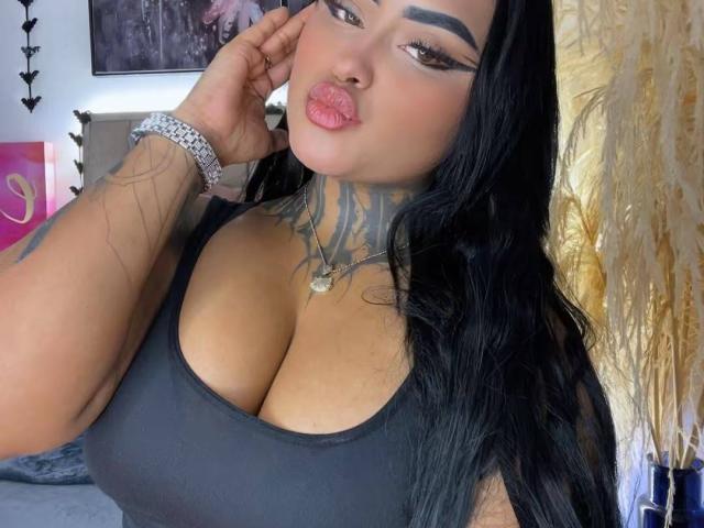 LairaHearvy ha un seno grande, un collo tatuato, labbra rosa carnose, capelli neri, e indossa una canotta nera con un braccialetto d'argento, guardando la fotocamera..