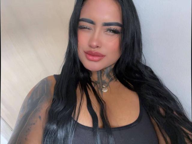 LairaHearvy heeft lang zwart haar, dikke wenkbrauwen en volle lippen met roze lippenstift. Ze draagt een zwarte tanktop, waardoor tatoeages op haar nek en arm zichtbaar zijn..