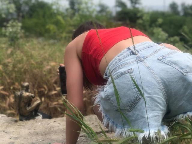 Milagres is bent over, wearing a red crop top and frayed denim shorts, showing her ass. She's holding a black camera in a grassy, dirt-filled area.