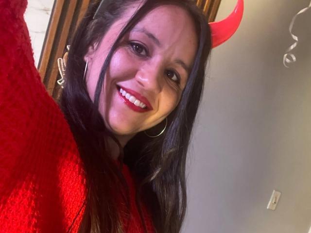 LilianaTavera with red devil horns, smiling, wearing a red sweater, hoop earrings, and red lipstick, holding up her phone for a selfie. Her tits are slightly visible.