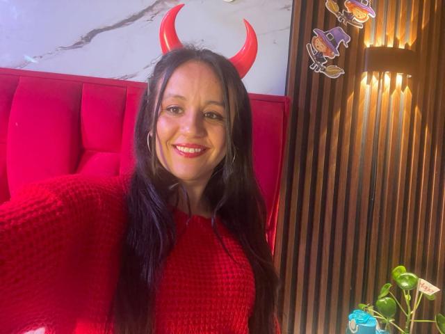LilianaTavera with red devil horns, long black hair, red textured sweater, smiling, large hoop earrings, sitting in red upholstered booth, wooden wall, green plant in bottom right.