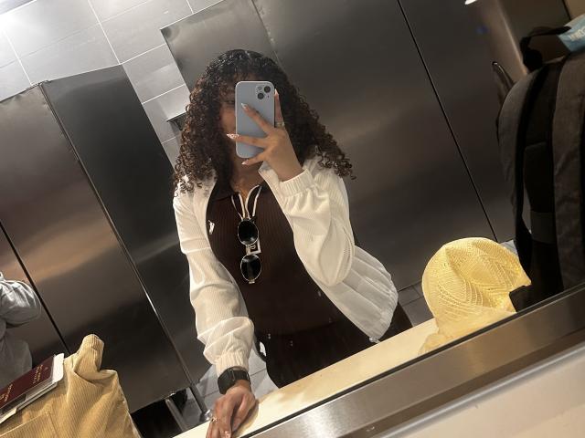 SofiaPassionne with curly hair, wearing a white jacket, brown dress, and sunglasses in a mirror selfie, holding a phone, standing in a bathroom, yellow cap on counter.