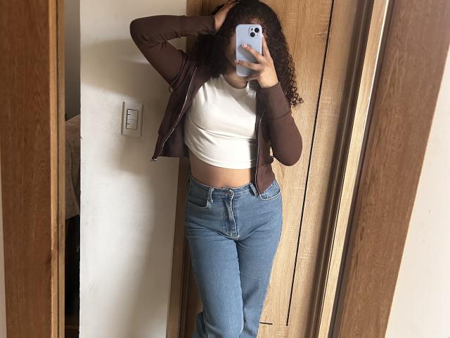 SofiaPassionne with curly hair, brown jacket, white crop top, and blue jeans, taking a mirror selfie, showing her midriff and holding a phone with a blue case.