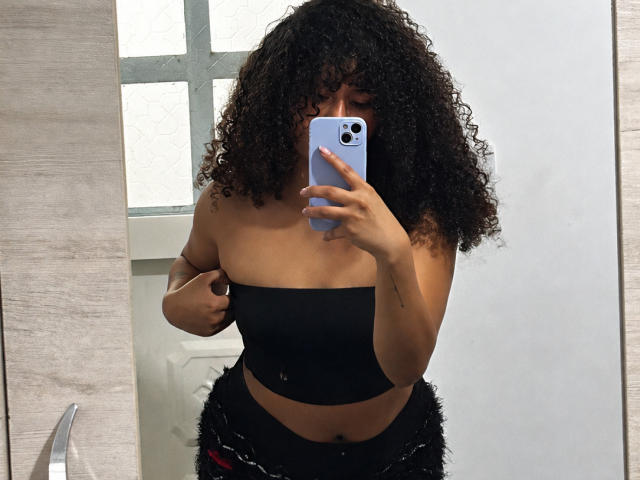 SofiaPassionne with curly black hair, holding a blue phone, wearing a black strapless top, showing her small tits, and black furry pants, taking a mirror selfie.