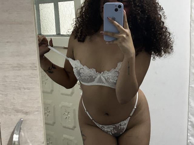 SofiaPassionne with curly hair, wearing white lace bra and thong, pulling bra strap down, holding phone in mirror selfie, showing her tits and pussy. Tanned skin, tattoos.