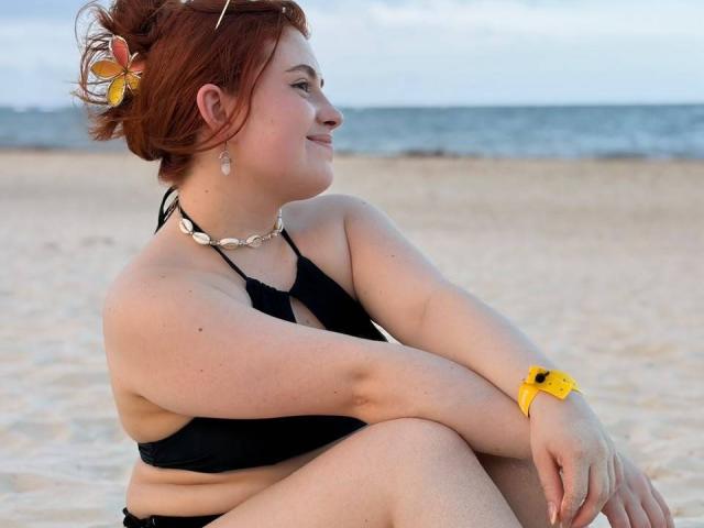 VicluX, a curvy redhead with small tits, sits on a beach in a black bikini, wearing a yellow watch, shell necklace, and yellow hair clip, smiling to the side.