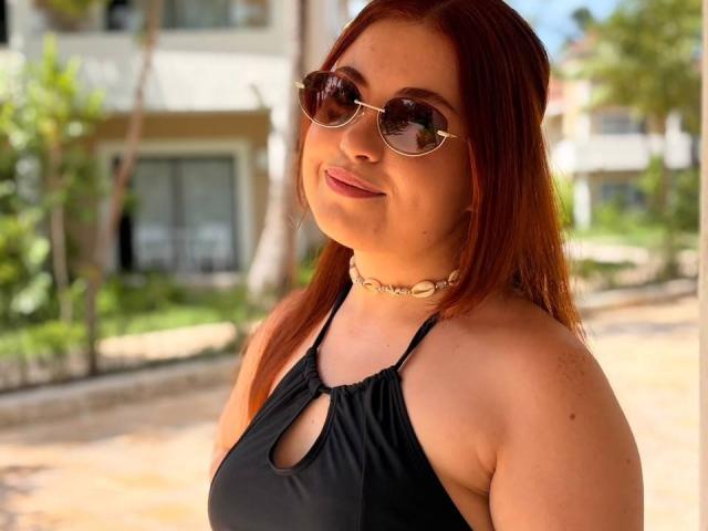 VicluX has red hair, wears round sunglasses, a black halter top with a cutout, and a shell necklace, smiling with a slight tan, in a sunny outdoor setting.