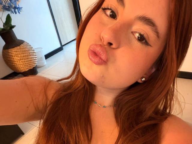 VicluX, com lábios rosados, olhos castanhos e cabelos ruivos longos, usando um colar delicado, exibindo seus ombros nus e pequenos brincos, em um selfie com iluminação quente..