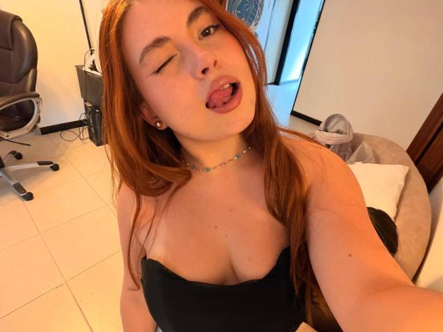 VicluX with red hair, winking and sticking out tongue, wearing black strapless top, showing cleavage, necklace, and small earrings, in a room with office chair and fan.