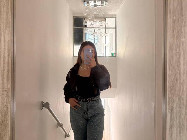 VicluX has long brown hair, wearing a black top, black jacket, and high-waisted gray pants with a studded belt. She's holding a phone, covering her face.