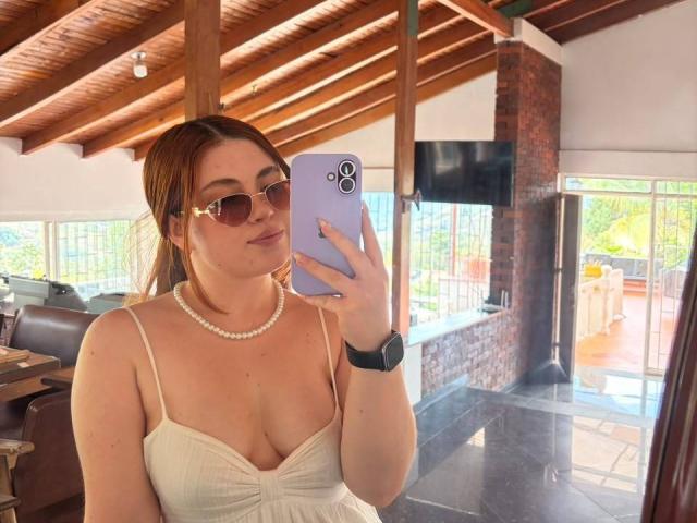 VicluX with glasses, pearl necklace, and white dress, holding a purple phone, showing her tits in a mirror selfie, wooden ceiling, brick wall, and a black watch.