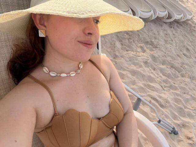 VicluX, com pele clara, cabelo ruivo, vestindo um top de biquíni bege, colar de conchas e chapéu branco, relaxando em uma cadeira de praia, mostrando seus seios..