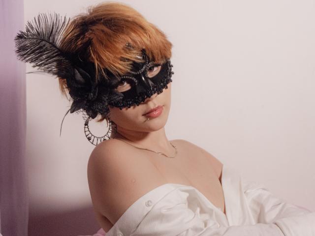 ChloeeWilliams with orange hair, black lace mask, feather, and hoop earrings, wearing an off-shoulder white shirt, looking seductive with a slight smile, showcasing her tits.