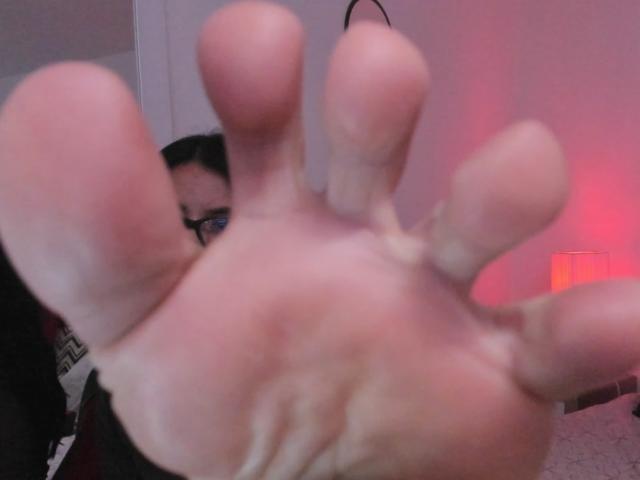 ZarayFeet's hand is in the foreground with slightly dirty fingers, showing a close-up of their boofing, slightly blurred face peeks from behind, wearing black glasses.