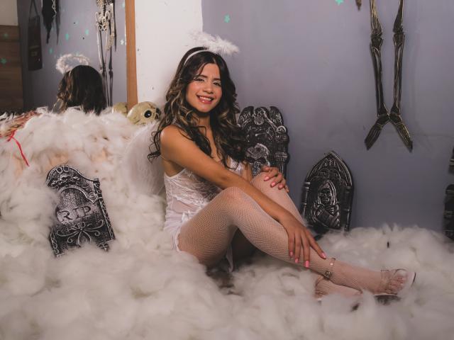 Sheyla Spooner, con cabello castaño rizado, alas de ángel blancas, lencería de encaje, medias de red, uñas rosas, sentada en una nube blanca esponjosa, sonriendo, con las piernas cruzadas, un halo blanco, almohadas decorativas negras..