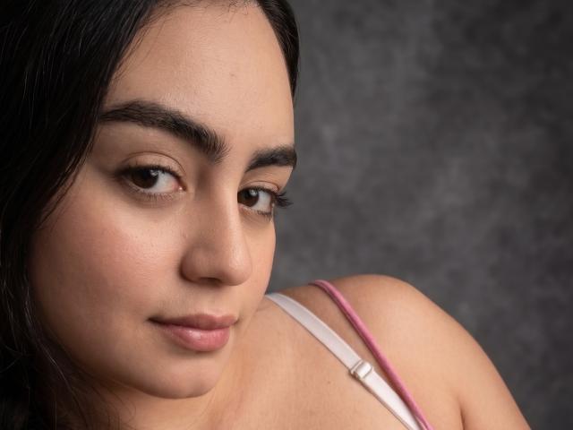 Daphne Calloway heeft een schattig gezicht met donkerbruine ogen, dikke wenkbrauwen en roze lippenstift. Ze draagt een roze bandje en haar schouder is zichtbaar. Zachte glimlach..