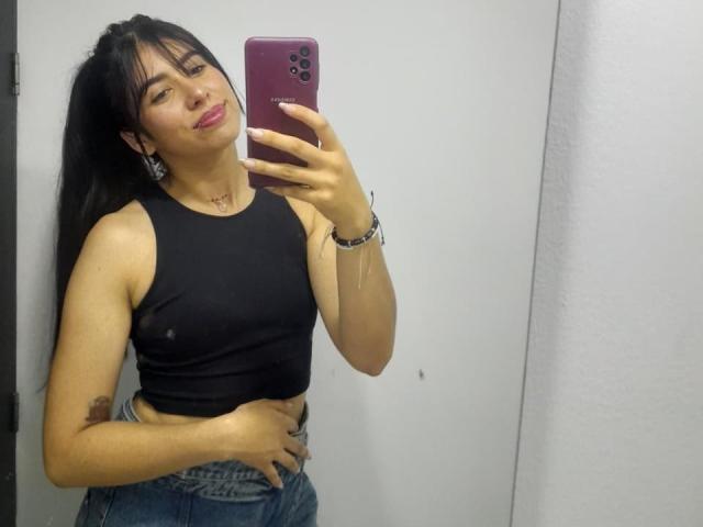 MilusSky with long black hair, wearing a black sleeveless top showing small nipples and blue jeans, taking a mirror selfie with a pink phone. Her hand's on her belly.