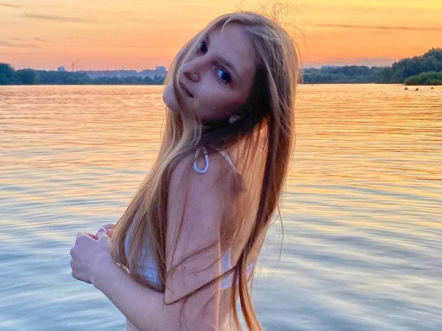 CathyBolyard with long blonde hair, looking over shoulder, wearing a white top, standing in water at sunset, holding her hair, showing her tits slightly, water reflection.