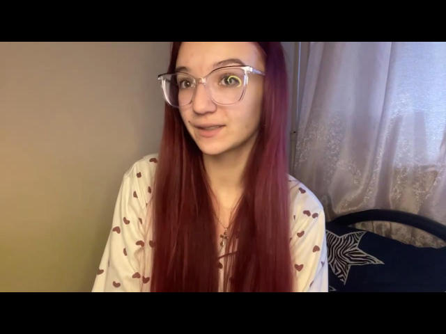 ThelmaSeilhamer with red hair, glasses, and a white shirt with heart patterns, has a cute, slightly nervous smile, showing her green eyes and a small nose.