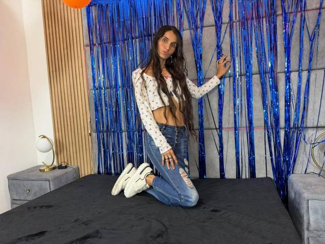 Tiffanny Conors è inginocchiata su un letto, indossa una canotta bianca a maniche lunghe, jeans blu strappati e scarpe da ginnastica bianche, con lunghi capelli castani e una figura snella e sensuale..