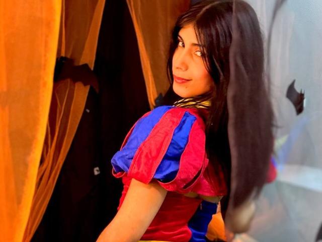 ValeryTranss with long dark hair, looking over shoulder, wearing a red and blue dress with puffy sleeves, standing by a brown curtain, smiling subtly.