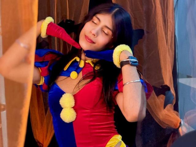 ValeryTranss with dark hair, eyes closed, wearing a red and blue jester outfit with yellow pom-poms, red gloves, and a smartwatch, smiling softly, holding her neck.
