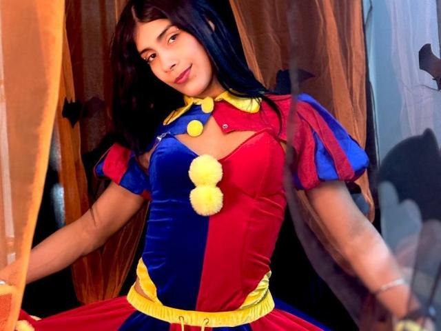 ValeryTranss with dark hair, wearing a red and blue velvet costume with yellow pom-poms, smiling, standing in a room with brown curtains, showing her small tits.
