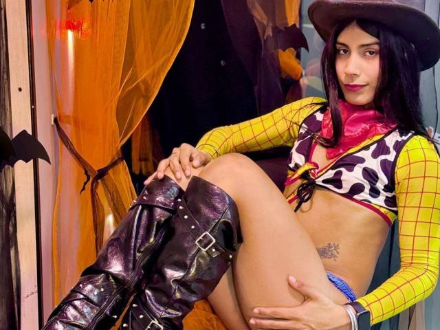ValeryTranss is wearing a cow print top, yellow checkered sleeves, red bandana, black cowboy hat, and shiny black boots, sitting with legs apart, showing a tattoo on her right hip.