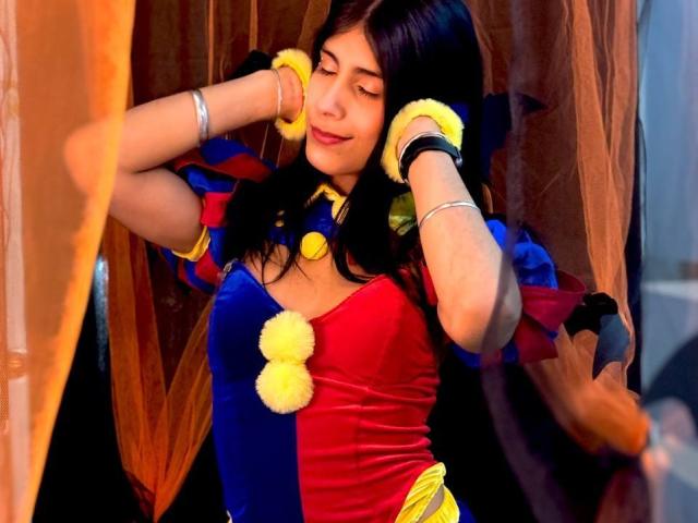 ValeryTranss with dark hair, wearing a blue and red velvet dress with yellow pom-poms, has her hands on her ears, smiling with closed eyes, and wearing yellow wristbands.