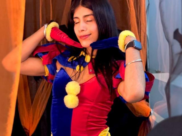 ValeryTranss, with long black hair, wearing a red and blue jester costume with yellow pom-poms, yellow gloves, and a black watch, smiling with eyes closed.