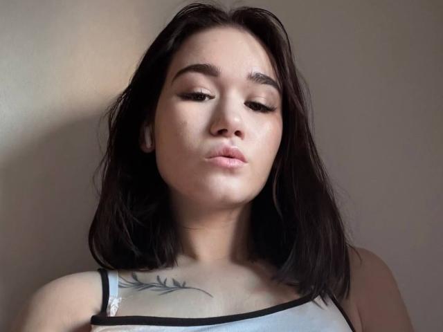 MaryanWelshans with dark brown hair, pouty lips, and a small tattoo on her right chest, wearing a white tank top with black straps. Her eyes are half-closed.