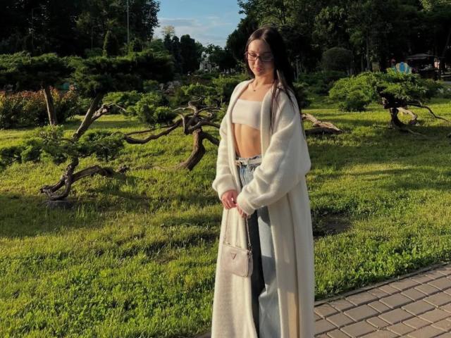 JaynaFeeler se tient dans un parc verdoyant, vêtue d'un débardeur blanc, d'un jean taille haute et d'un long manteau blanc, tenant un sac beige, avec de longs cheveux noirs et des lunettes..