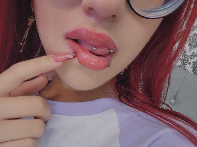 CherryBuchel's face with red hair, wearing glasses, pink glossy lips with a labret piercing, and a finger touching her lip. She's wearing a purple and white shirt.