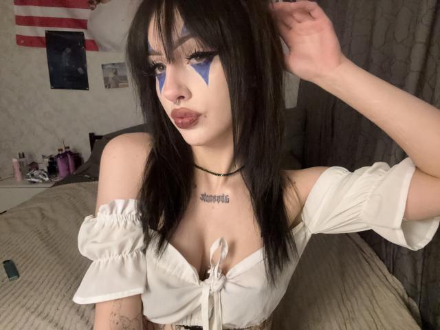 SimonneBrustkern with black hair, blue face paint, white off-shoulder top, showing tits, tattoo on chest, pouty lips, hand in hair, choker, in a bedroom setting.