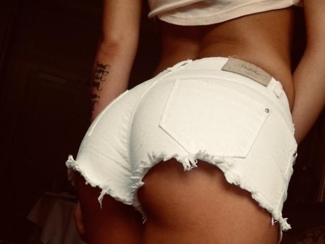 A BlackAngeel está usando shorts jeans brancos, desgastados e justos, que abraçam suas nádegas arredondadas e bronzeadas. Ela veste uma blusa branca, e seu braço tatuado está parcialmente visível..