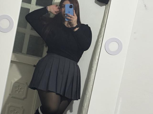 VenusxMusa trägt ein schwarzes Oberteil, einen grauen, gefalteten Rock, schwarze Strumpfhosen, hält ein blaues Telefon, Selfiemotiv, Arm erhoben, langes, braunes Haar, weiße Wand, reflektierender Spiegel, leicht sichtiger Hintern..
