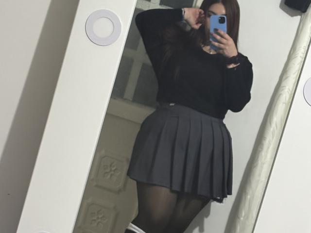 VenusxMusa con lunghi capelli castani, una maglietta nera a maniche lunghe, una gonna grigia plissettata, collant neri, che tiene in mano un telefono blu, un selfie allo specchio, braccio destro sollevato, una porta bianca visibile sullo sfondo..